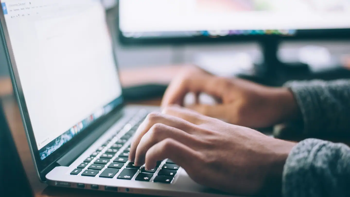 Hands typing on a laptop keyboard with a second monitor showing a webpage.