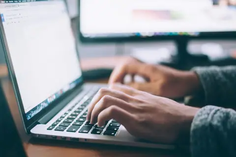 Hands typing on a laptop keyboard with a second monitor showing a webpage.
