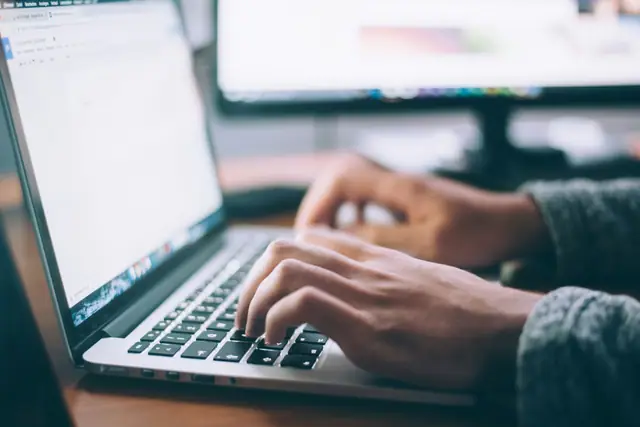 Hands typing on a laptop keyboard with a second monitor showing a webpage.