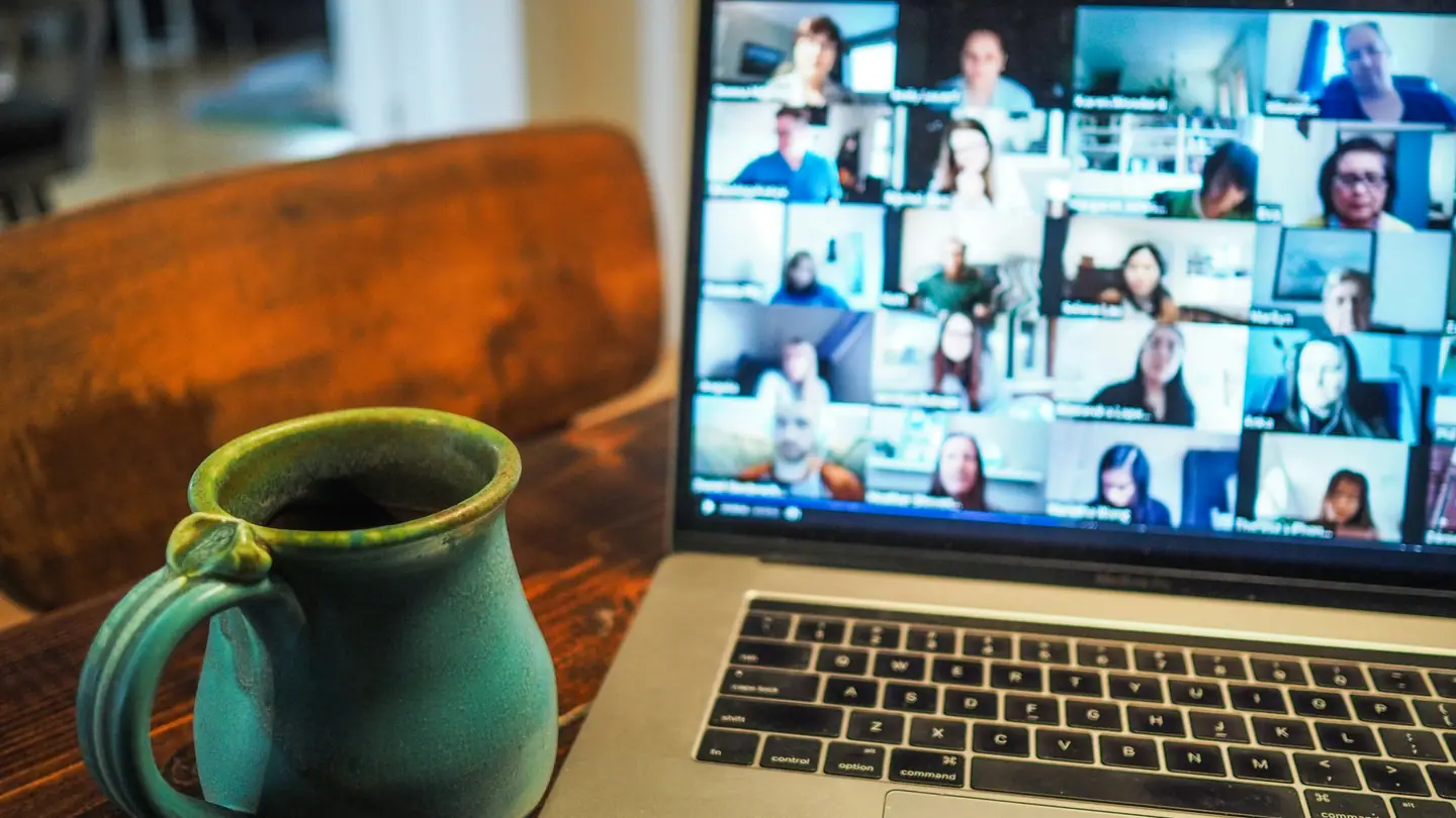 A laptop on a wooden table showing a grid of video-call participants, a mug beside it.