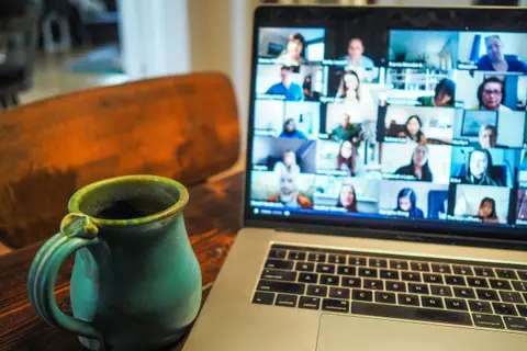 A laptop on a wooden table showing a grid of video-call participants, a mug beside it.