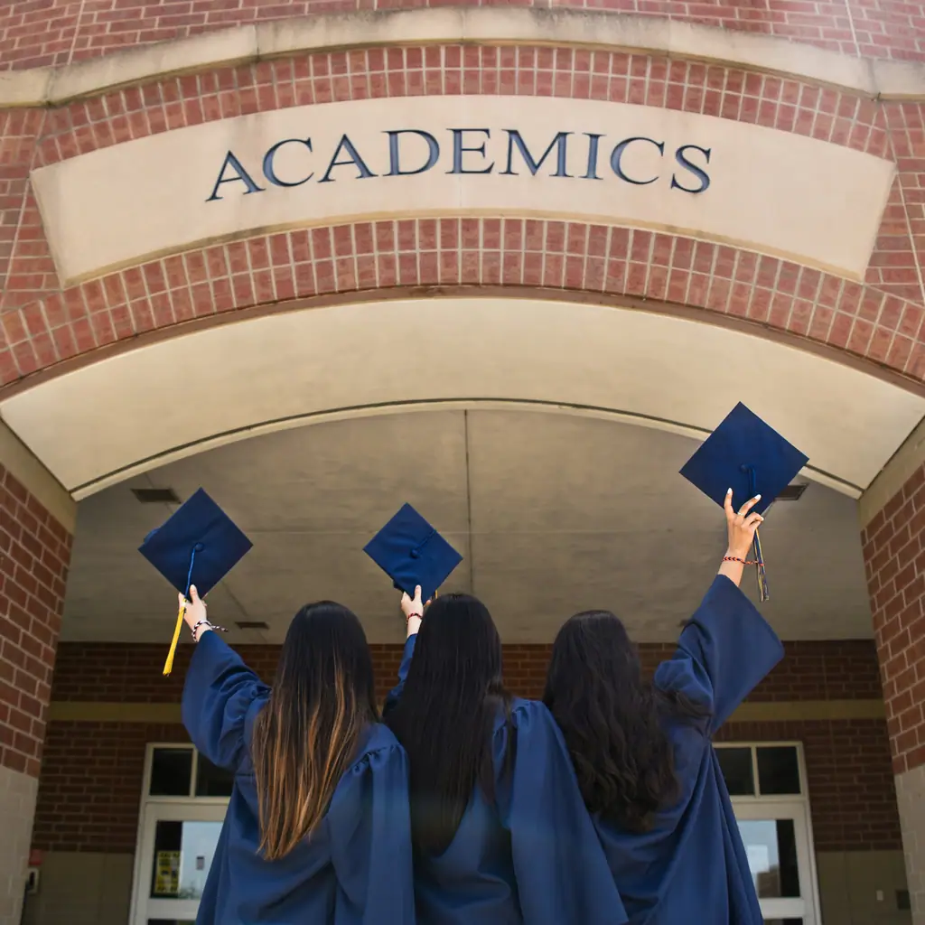 Three graduates in navy gowns raising their mortarboards beneath a brick archway marked “Academics”.