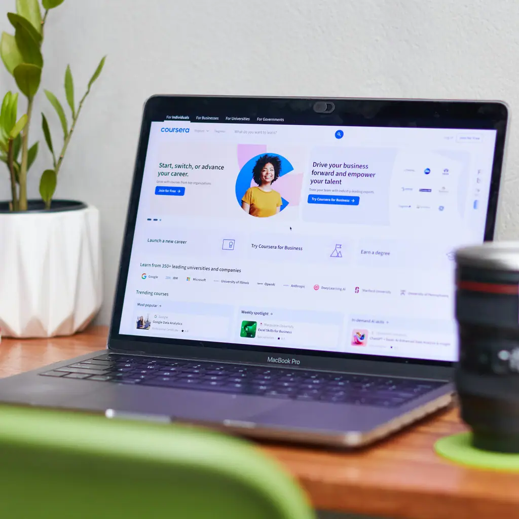 A laptop on a wooden desk showing an online course platform’s homepage, beside a potted plant and a notebook.