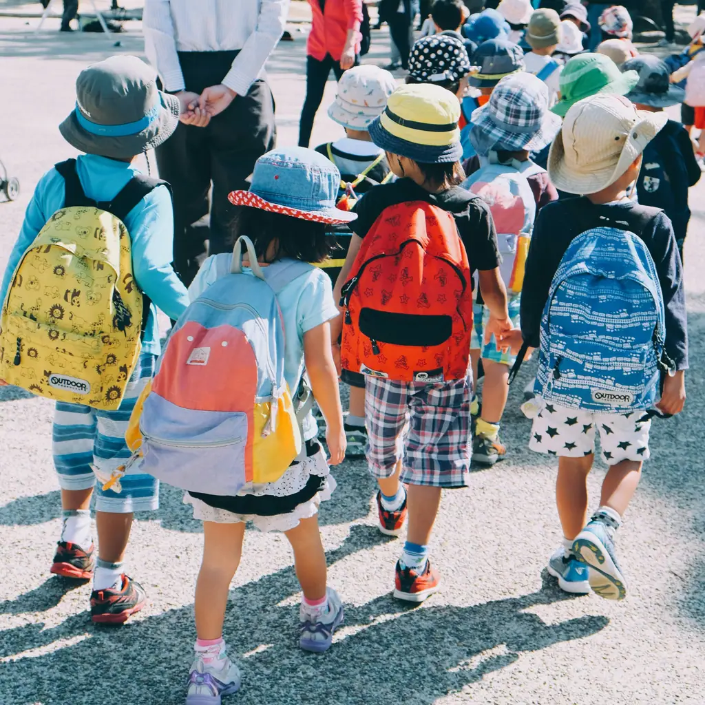 Young pupils in sun hats and colourful backpacks walking together across a school yard.